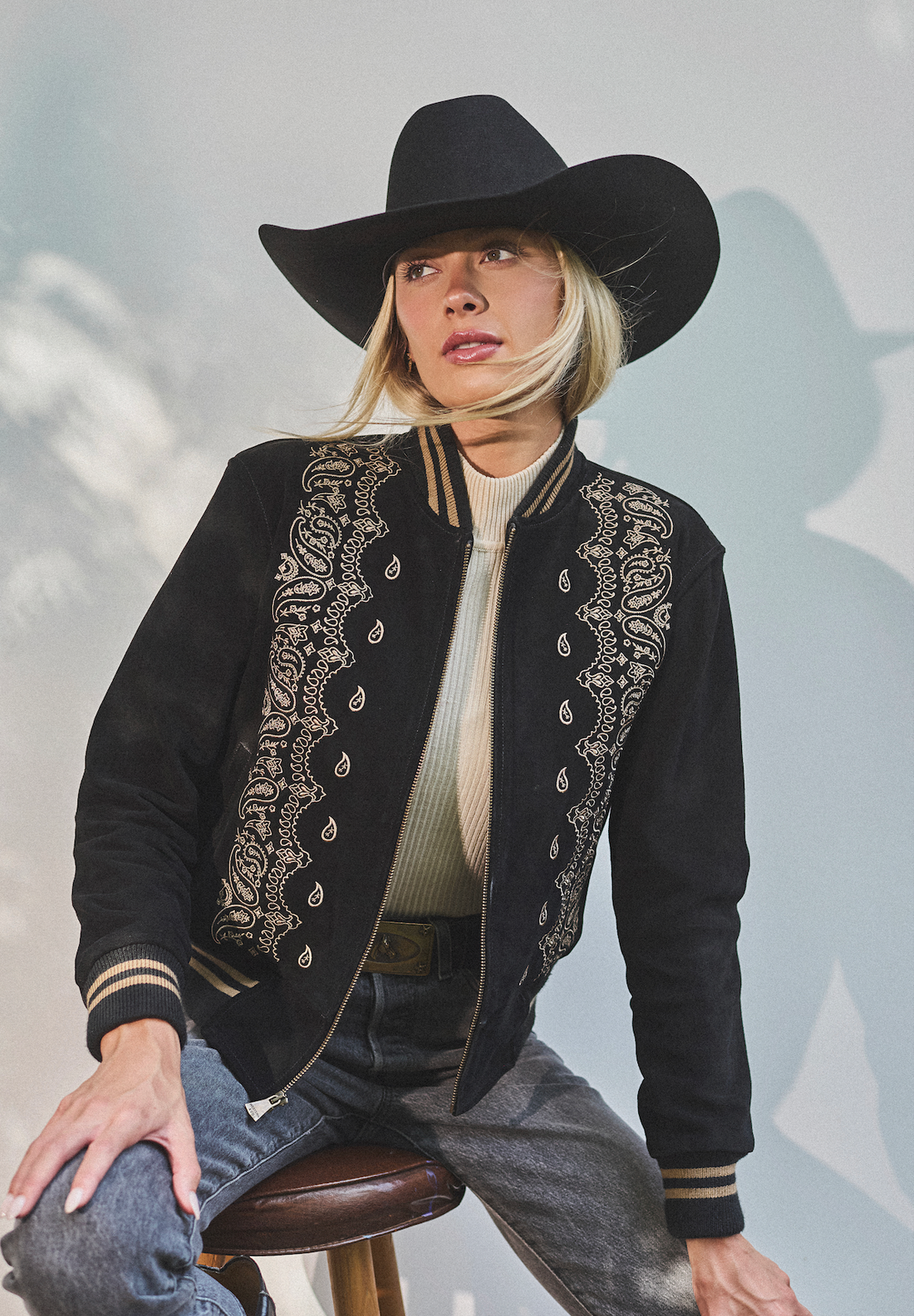 Woman in a cowboy hat wearing black embroidered bomber jacket by Western & Co sitting on a stool against a light background 