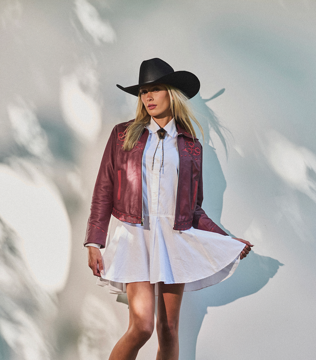 Woman in a cowboy hat, white dress wearing embroidered sangria jacket by Western & Co against a light background 