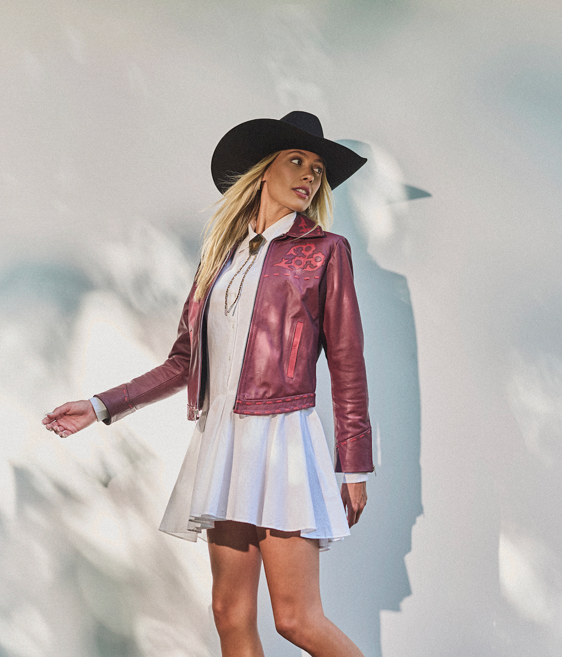 Woman in a cowboy hat, white dress wearing embroidered sangria jacket by Western & Co against a light background 