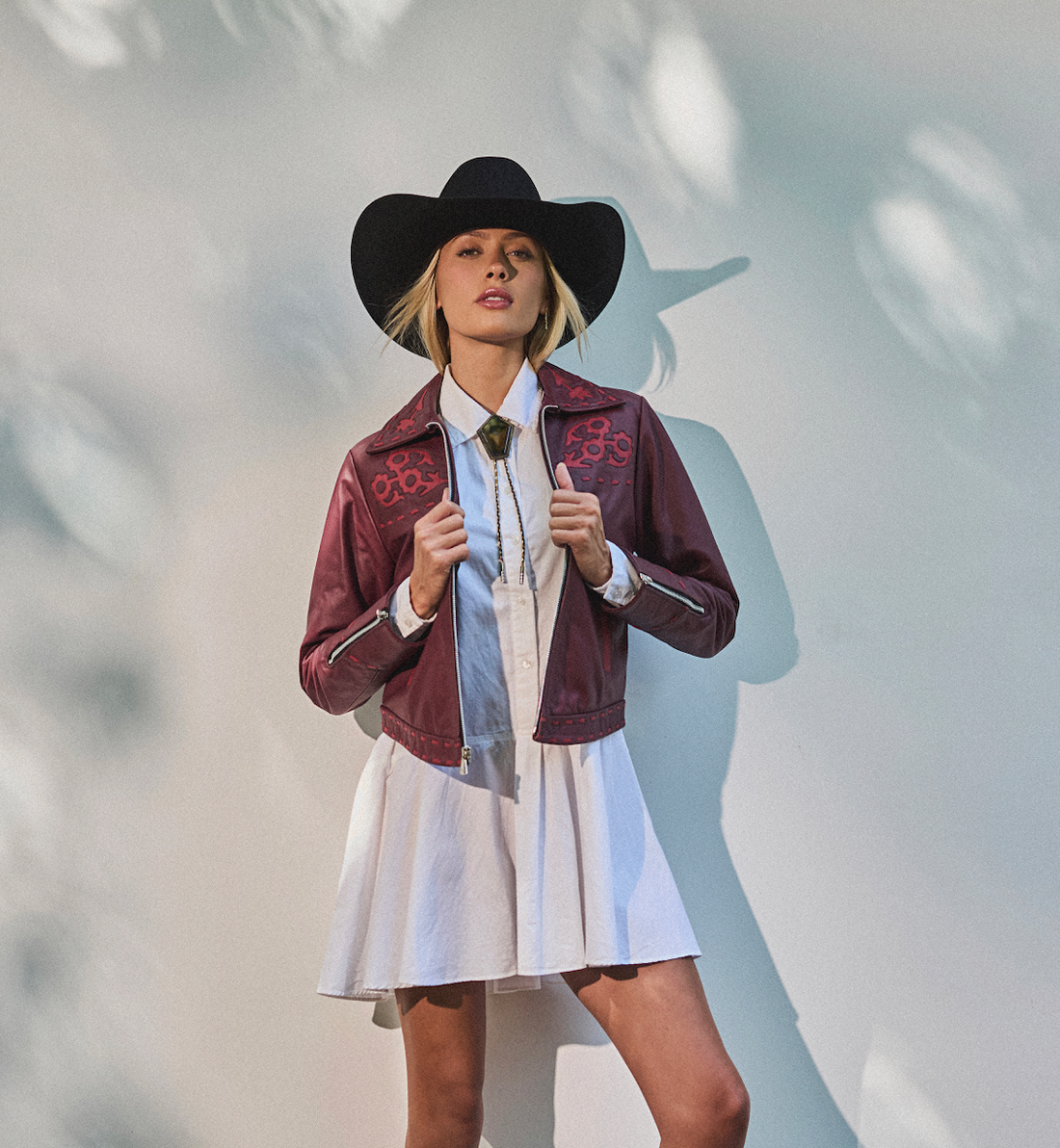 Woman in a cowboy hat, white dress wearing embroidered sangria jacket by Western & Co against a light background 